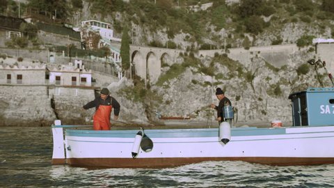 Two hardworking fishermen bring out nets for fishing sardines in the sea with view of the Amalfi Coast in the background. Wide shot on 8k helium RED camera.