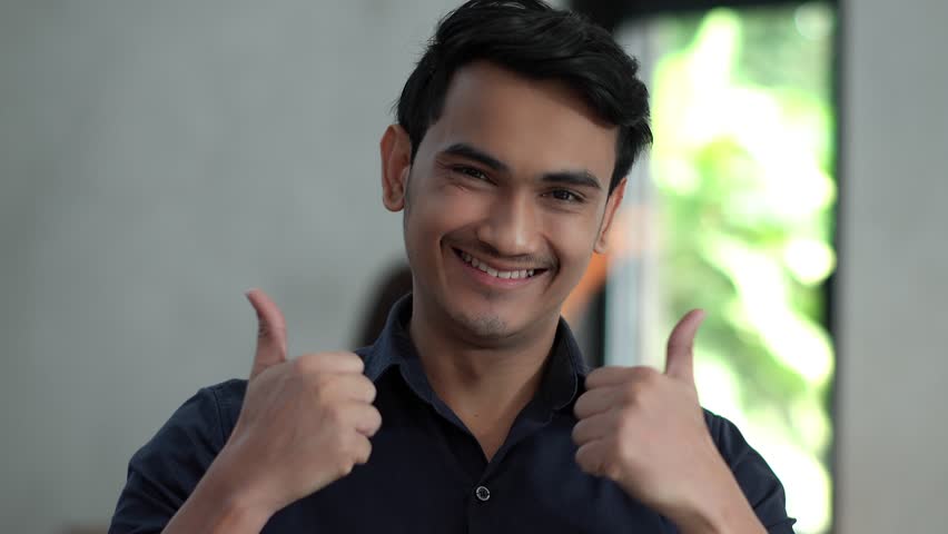 4K locked shot portrait of happy expressions of young handsome man standing looking at camera with smiling and thumbs up in cafe, emotion concept