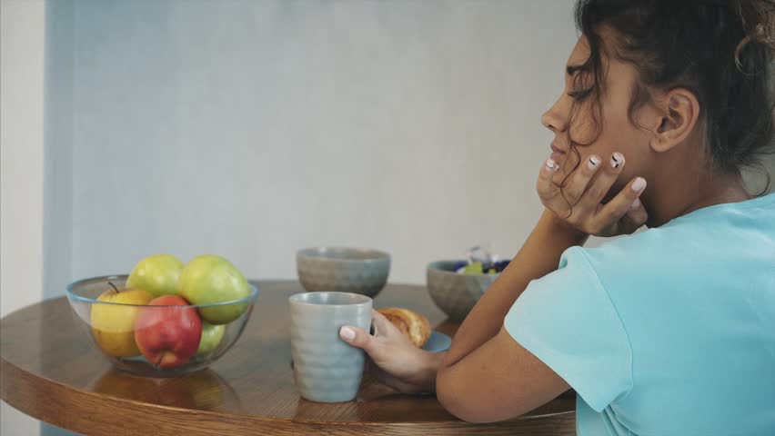 Beautiful young slender brunette with green apple in one hand and croissant in the other hand on background of vegetables on kitchen table making a difficult choice between healthy and harmful food.