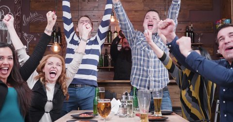 Group of friends are watching a sport game on TV in a vintage pub or restaurant. They all cheer when their favourite team scores