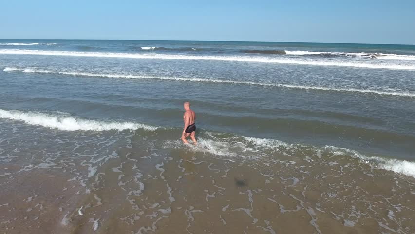 The athlete runs along the beach to the sea. A man of strong build runs along the beach to the sea. Athlete, European. Bright sun and clear sky.