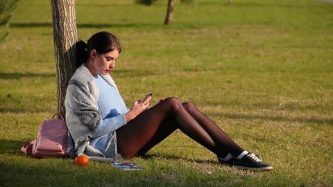 Cute brunette girl with beautiful long legs in shorts and sneakers sits on green grass near tree and looks up something in phone and eats red apple on sun day, next to her are fruits and pink backpack