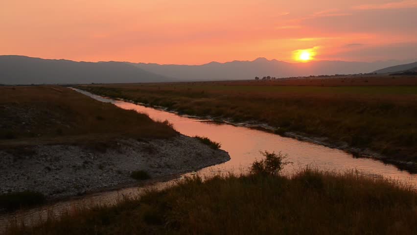 Red Sunset over rivers and green field with mountains in distance. Orange sunset over meadow and water canal. Beautiful countryside landscape. Summertime