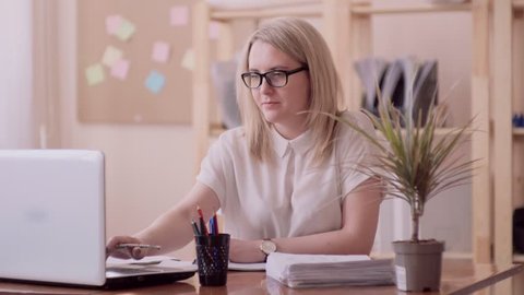 blonde girl of caucasian appearance in glasses right-handed in the office to the right of the flower sitting at the laptop checks the paper and writes notes with a pencil.