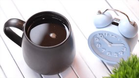 A mug of black coffee on a white wooden table with alarm clock. Selective focus. Tilt up footage.
