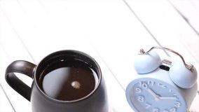 A mug of black coffee on a white wooden table with alarm clock. Selective focus. Tilt down footage.