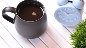  A mug of black coffee on a white wooden table with alarm clock. Selective focus. Panning to the right.