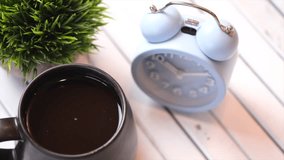  A mug of black coffee on a white wooden table with alarm clock. Selective focus. Panning to the right.