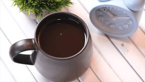 A mug of black coffee on a white wooden table with alarm clock. Selective focus. Panning to the left.