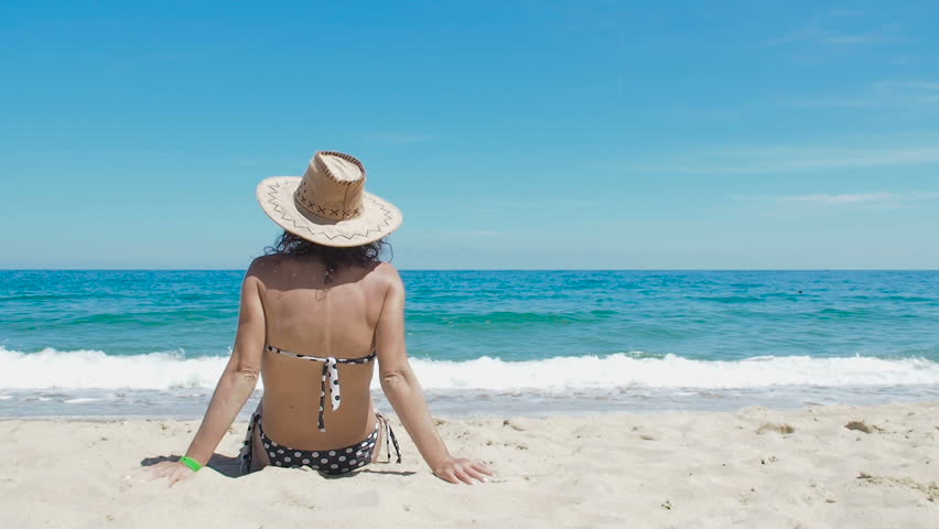 Happy woman on resort sitting on sand. Woman raises her hands up. Wearing hat. Clear sky and blue water.