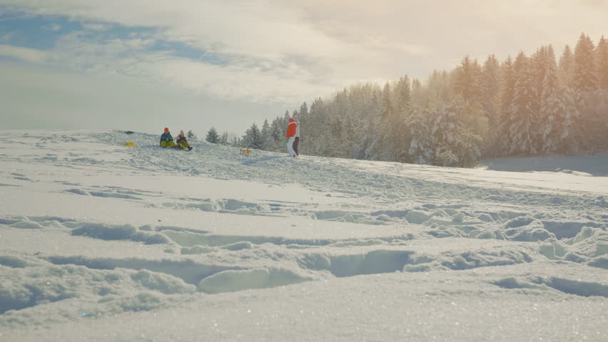 Two kids sledding down a hill while a teenager is chasing her dog