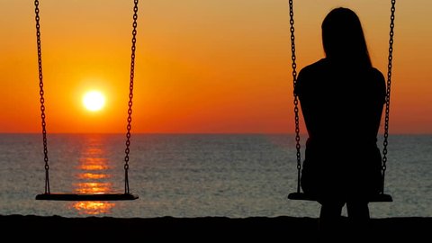 Back view silhouette of a sad girl swinging looking at side t an empty seat on the beach at sunset