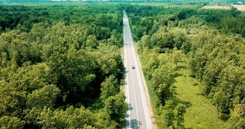 Wide shot aerial drone footage tracking a sedan car cruising on an empty road surrounded by a beautiful large forest and river.