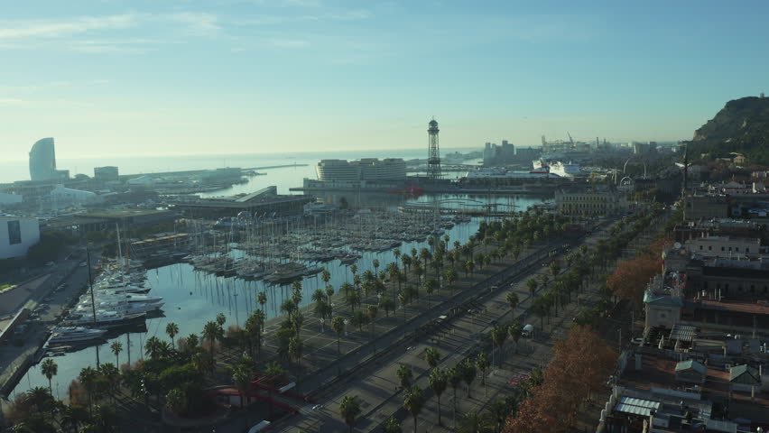 Aerial view of city port in Barcelona; parking for large luxury yachts in touristic capital of Catalonia; sunny day in Barcelonetta, sailboats, hotels, old buildinags illuminated by warm morning sun Royalty-Free Stock Footage #1023898336