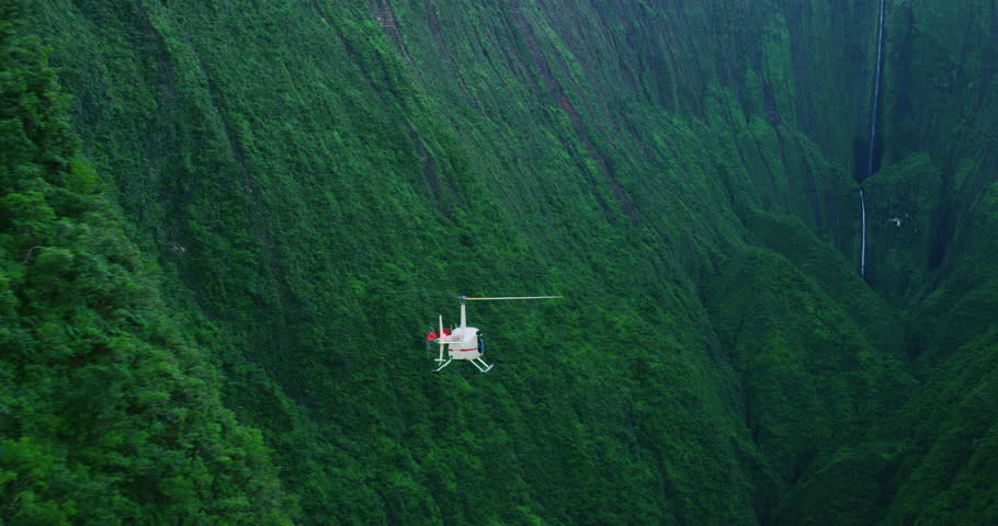 Aerial shot of helicopter approaching giant waterfall in lush green valley, epic helicopter adventure