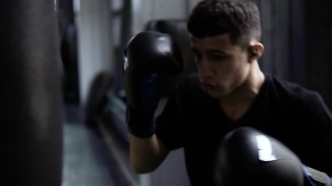 Handhelded footage of young boxer's training. Dark haired man hitting the boxing bag, hard kicks. Motivation in sport. Old style gym, daytime. Close up 庫存影片