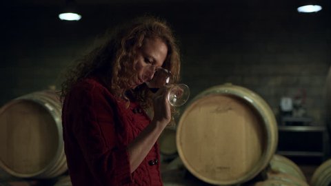 In a wine cellar, a woman takes a sample of red wine from a barrel into her glass and smells it. Medium shot on 8k helium RED camera.