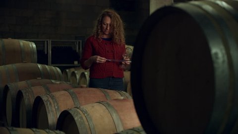 In a wine cellar, a woman takes a sample of red wine from a barrel into her glass and smells it. Medium shot on 8k helium RED camera.