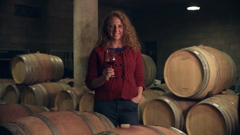 In a wine cellar, portrait of a woman woman holding a glass of red wine with barrels in the background. Medium shot on 8k helium RED camera.
