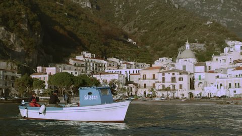 Two busy fishermen sailing in a boat along the ocean with a view of the Amalfi Coast in the background on a bright sunny day. Wide shot on 8k helium RED camera.
