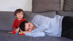 Funny little boy sitting on a sofa in red sweater near tired asleep father. Side view