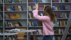 Rear view of female student in pink sweater and jeans dancing against bookshelves background. Blonde long-haired caucasian woman in bluetooth headphones making musical break in library