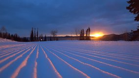 snowmobile rides on the track prepared snowgroomer with grooves, corduroy. red sunrise and splashes of snow in the background light. mountain forest and cloudy sky. stock video footage. Slow motion