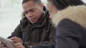 Afro-American middle-aged man in black jacket with fur hood discussing data with Afro-American young girl with braids, showing data at tablet. Communication lifestyle, wireless technology concept