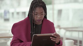 Afro-American attractive young girl with plump rose lips and braids in aviator eyeglasses wearing rose coat, sitting outside and working on tablet. Communication lifestyle, wireless technology concept