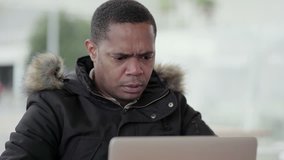 Afro-American middle-aged man in black jacket with fur hood working on laptop outside, finding out mistake, becoming angry and disappointed. Medium shot. Work, wireless technology concept