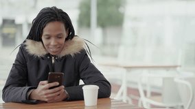Afro-American happy young girl with braids in grey jacket with fur hood sitting outside, texting on phone with friend, turning head to camera and smiling. Leisure, wireless technology concept