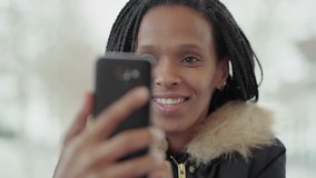 Afro-American cheerful young girl with braids in grey jacket with fur hood sitting outside, having video chat on phone, talking with friend. Close up shot. Leisure, wireless technology concept