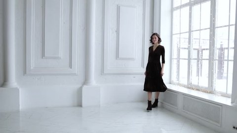 elegant girl in black dress smiling and posing in white room with big Windows, slow motion स्टॉक वीडियो