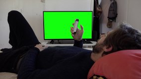 Using Smartphone Lying In Bed Watching TV Green Screen Television. Young man relaxed at home using a smartphone and watching television green screen. Shot behind models shoulders