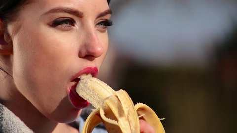 Close-up attractive sexy young brunette woman with beautiful eyes and red lips eats banana on sunny day. Spring beriberi, vitamins in fruits.