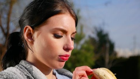 Slow motion, close-up attractive sexy young brunette woman with beautiful eyes and red lips eats banana on sunny day. Spring beriberi, vitamins in fruits.