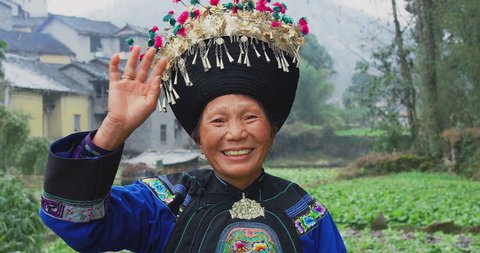 Portrait of old miao woman waving hello,  in laojiazhai village, dressed it traditional cultural costume. Minority people in china villages. Slow motion, red cinema camera, hand held. 