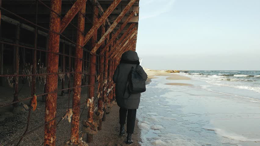 girl walking along the seashore near metal rusty structures
