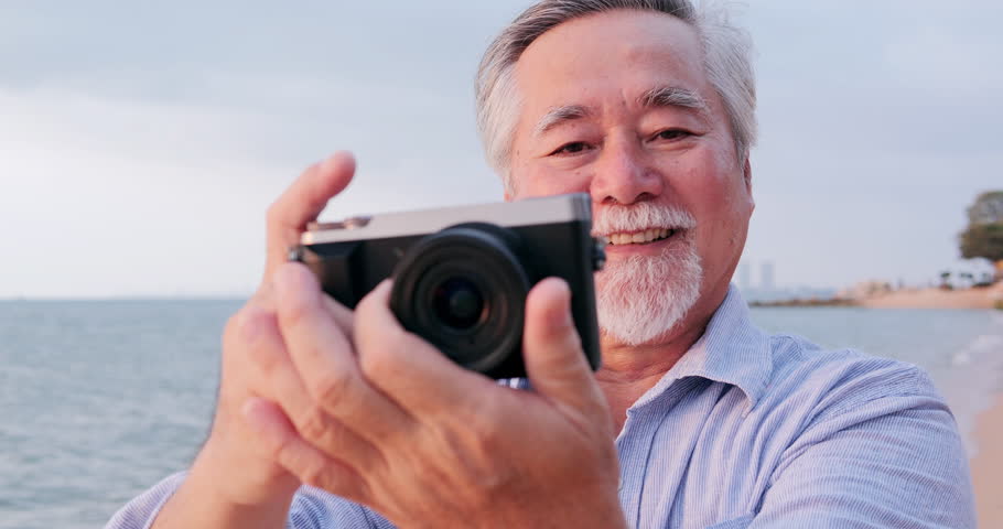 Senior asian man using camera on the beach with happy emotion. People with lifestyle, retirement and technology concept.