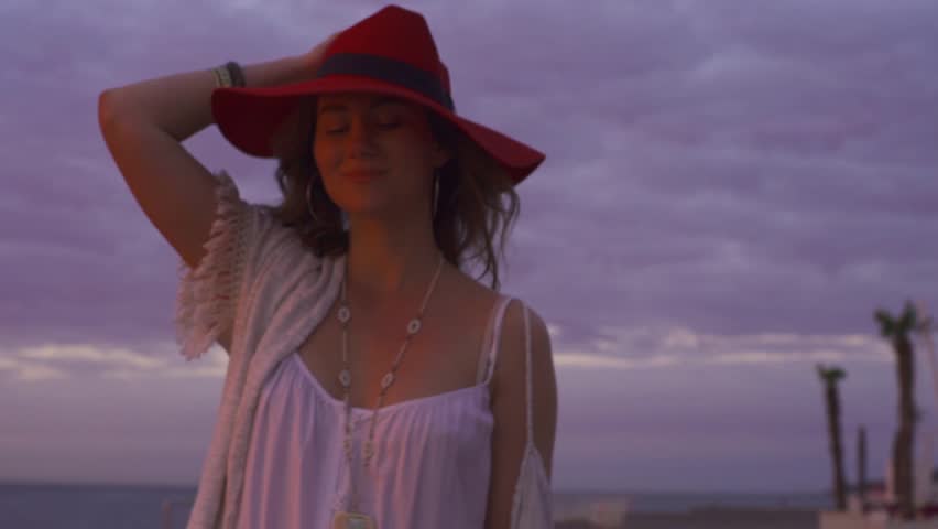 young attractive woman walking at sea on sunrise in white boho dress wearing red hat, relaxing on vacation
