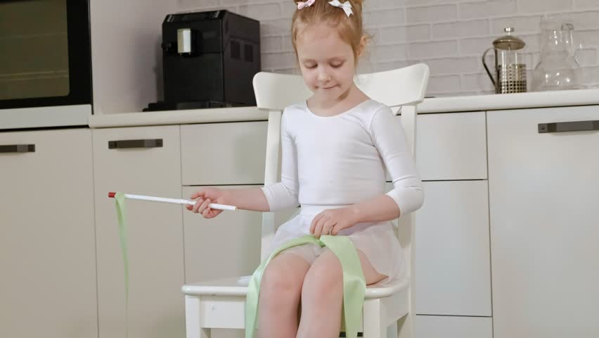 A happy little girl with blond hair in a white gymnastic swimsuit with a skirt trains, dances at home on a chair with a green ribbon for rhythmic gymnastics in a white room on a green carpet, jumps