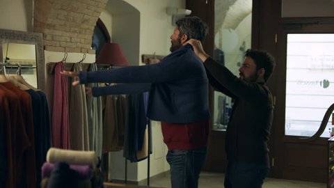 In a clothing store, an Italian man tries on a sweater and looks in the mirror as a store clerk helps him. Medium shot on 8k helium RED camera.