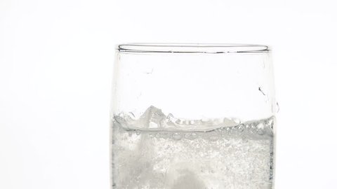 Close up dropping ice cubes in glass of pure clear sparkling water with gas over white background, low angle side view, slow motion
