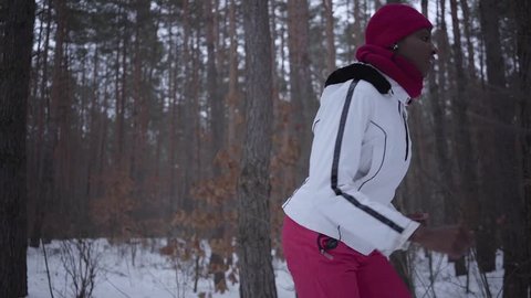 Scared African american young woman dressed warm wearing a red hat, scarf and white jacket running through the snowy forest. The girl constantly looks back running away from the pursuer. Slow motion.
