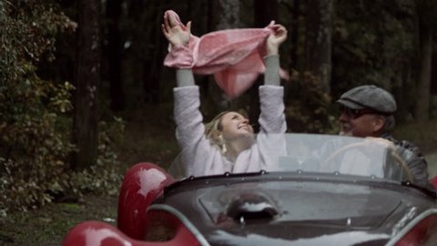 Older boyfriend driving in a convertible sports car with his younger girlfriend, as she throws her scarf in the air happily. Medium shot on 8k helium RED camera.