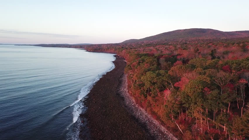 Ascending shot over a coast line in Maine. Fall colors, vast forest lines the edge of the land, rocky shore, then the atlantic ocean to the left. Taken at sunrise this shot is a gorgeous reveal.