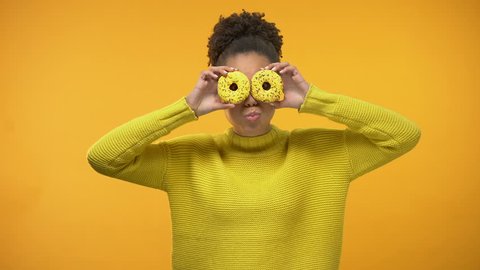 Cheerful Afro-American girl closing eyes with donuts, having fun, background