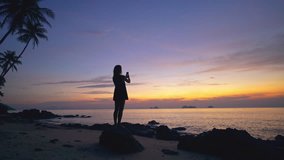 Young woman with smart phone in hand is standing on a rock by the sea on a sunset background and shoots a video 