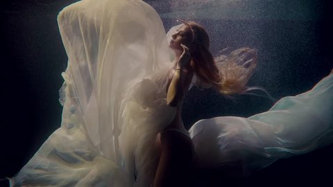 Graceful young woman is floating underwater in light beams. She is naked, covering breasts by white silk cloth, relaxing and posing