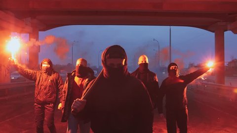 Group of young men in balaclavas with red burning signal flare walking on the road under the bridge, slow motion